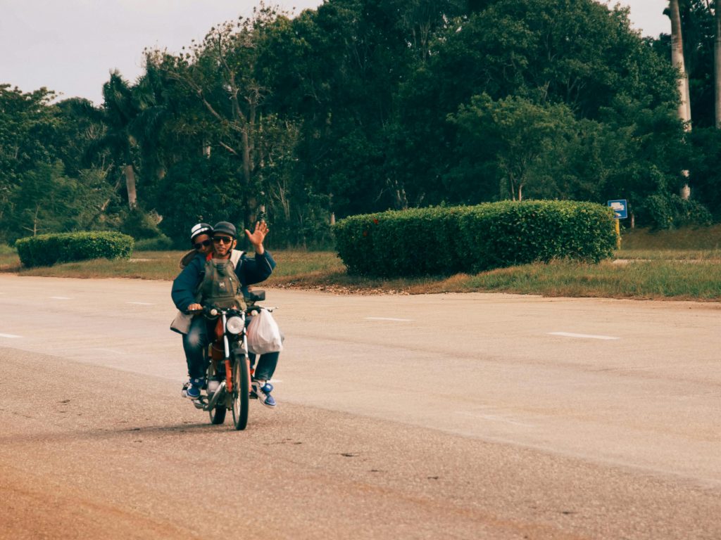 Two adults riding a motorcycle on a rural road, one waving joyfully.