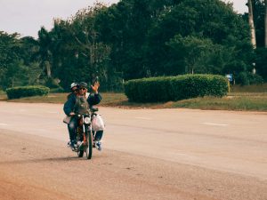 Two adults riding a motorcycle on a rural road, one waving joyfully.